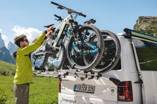 A man with a green functional jacket and sunglasses mounted an e-bike on the bicycle carrier of the CamperBoys Camper during his bicycle holiday, which stands in a mountain landscape